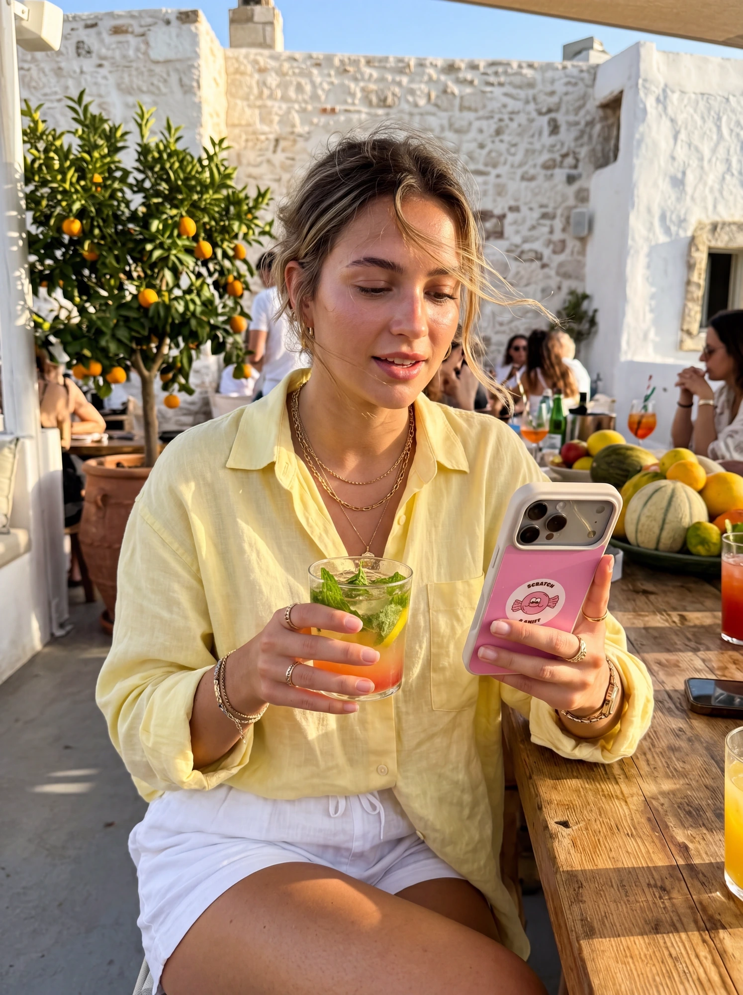 Woman in a linen yellow shirt at a Mediterranean rooftop cafe holding a mojito and a pink phone case