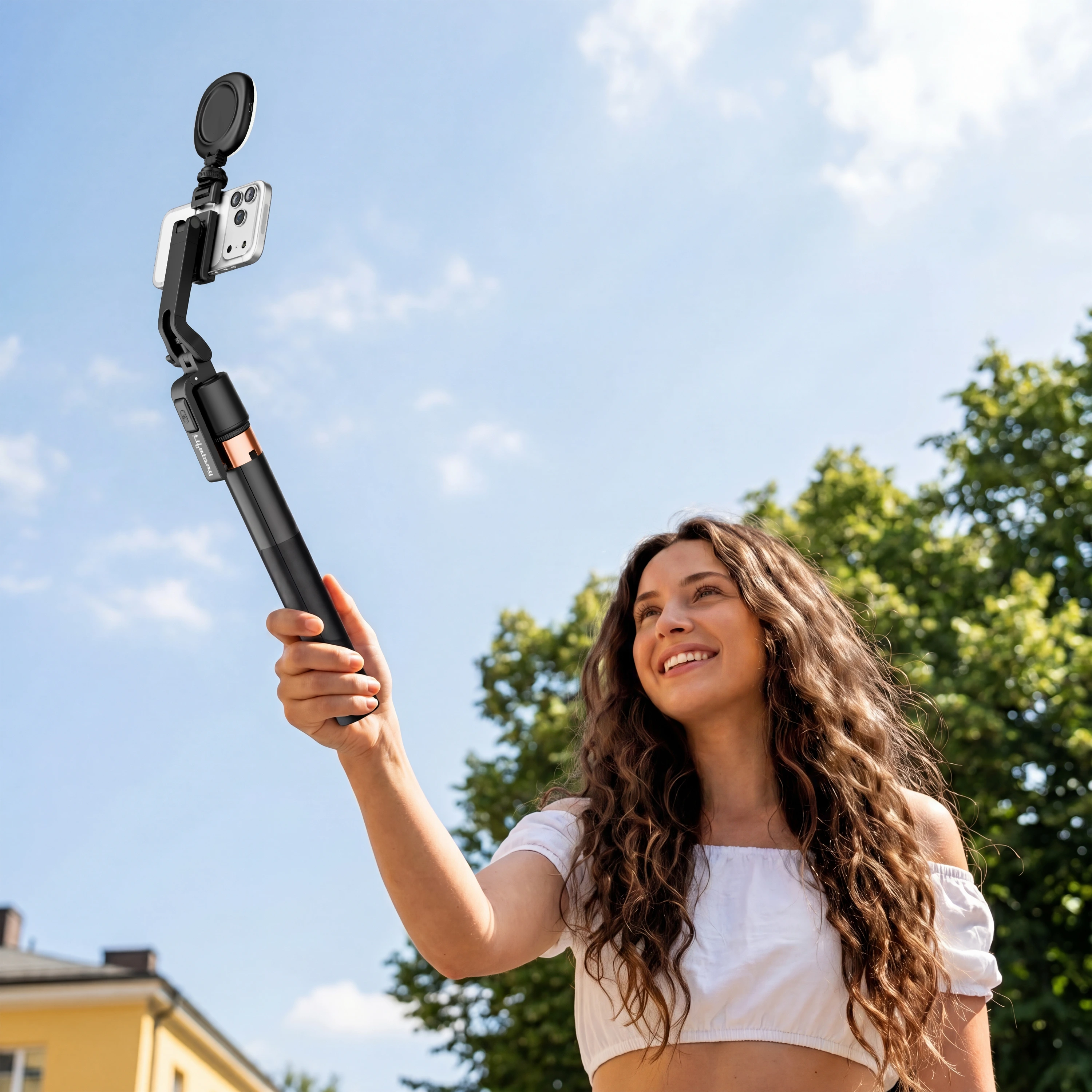 Woman with curly hair holding a black handheld gimbal-selfie-stick against a bright summer sky